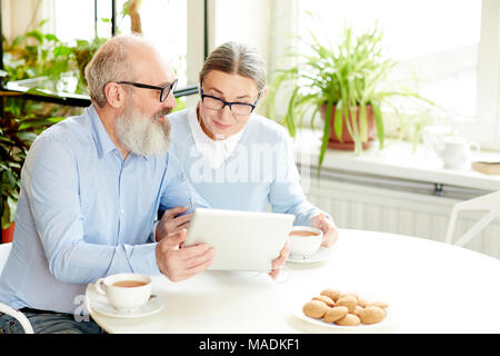 Couple contemporain avec tablet et écouteurs de parler à travers le vidéo-chat assis dans le café par tasse de thé avec les cookies Banque D'Images