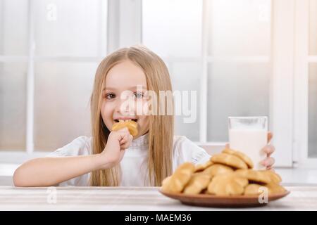 Petite fille a bien des biscuits et du lait sur le dîner. Elle prend de grandes plaques à motifs des gâteaux de Noël et des boissons de lait en verre transparent.Ils demeurent le tableau,recouvertes de nappe. Banque D'Images