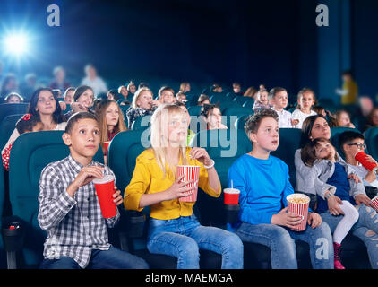 Façade de peu quitté les enfants regardant bon film en salle de cinéma. Les garçons et filles semblent très émus et heureux. camarades portant des vêtements colorés,eating popcorn, boire des boissons gazeuses. Banque D'Images