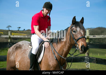 Beau mâle Horse Rider à cheval avec un casque, un pantalon blanc, des bottes noires et rouges sur fond de ciel bleu polo en paddock Banque D'Images