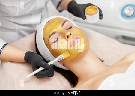 Spécialiste de la cosmétologie mains dorer à l'aide de masque du visage, ce pinceau peau hydratée et le visage radieux et la peau. Attractive brunette relaxing wi Banque D'Images