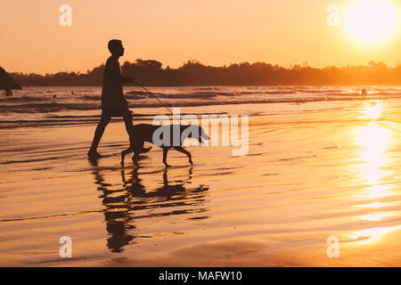 Silhouette d'un homme marchant avec un chien en laisse le long de la plage au coucher du soleil. Banque D'Images