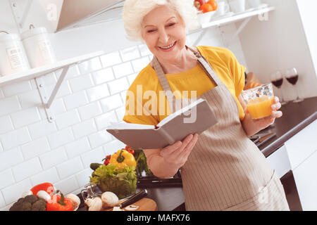 Bien que les croissants sont cuits, grand-mère boit du jus d'orange et lit son livre préféré. Piscine, studio tournage, cuisine intérieur Banque D'Images