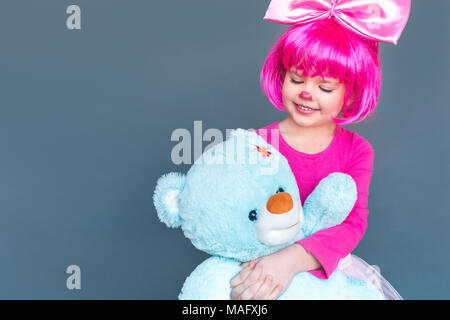 Bonheur petite fille en rose perruque , est titulaire d'ours en peluche, serrant et souriant à pleines dents. Studio shot, isolé sur fond gris Banque D'Images