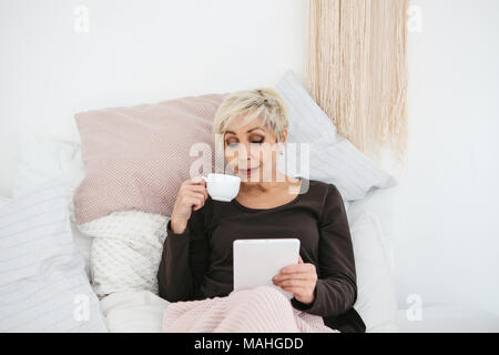 Une vieille femme au lit de boire le café du matin utilise une tablette pour lire les communiqués ou discuter avec vos amis sur un réseau social. La génération des personnes âgées et nouvelles Banque D'Images