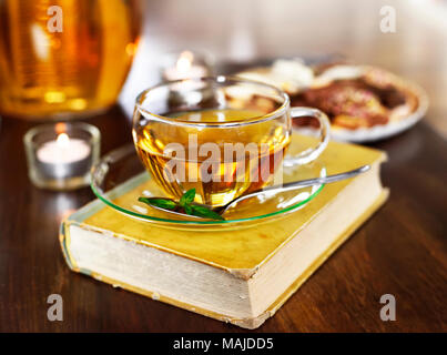 L'heure du thé théière en verre avec scène et une tasse de thé, livre ancien et d'un bain de vapeur. L'heure du thé ou idyllique scène relaxation avec les cookies et des bougies. Banque D'Images