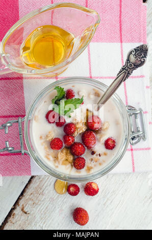 Yaourt au lait frais avec les fraises et granola servi dans un bocal en verre avec les feuilles de menthe et le miel sur la table en bois blanc, rouge à rayures serviette. Banque D'Images