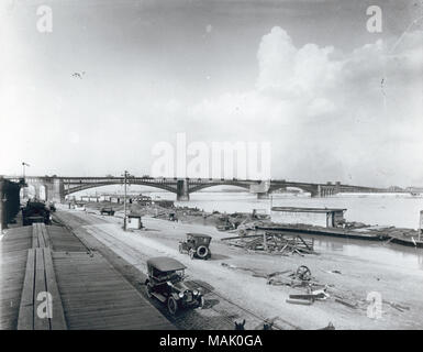 L'horizontale, 1970 Photographie noir et blanc de la rivière avec le pont avant d'Eads dans l'arrière-plan. Les voitures sont garées sur le bord de la rivière et le bateau Mary S. Blees est amarré le long de la banque. Des piles de bois sont cassées sont empilés le long de la banque et des ponts pour piétons sont présents dans la distance. Titre : Saint Louis de digue, vue nord vers Pont d'Eads avec le bateau du quai, du vapeur Mary S. Blees. . 1915. Banque D'Images
