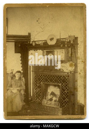 Photographie sépia, verticale montrant une femme assise à la gauche d'une cheminée. Il y a de nombreux articles sur le manteau dont un réveil, de la direction générale dans un vase, une assiette, et deux chandeliers. Titre : Femme assise à côté d'une cheminée allumée. . Vers 1880. Banque D'Images