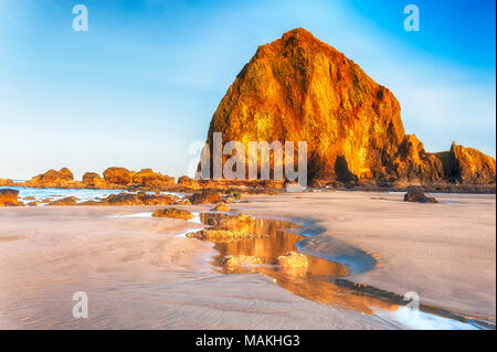 Le soleil levant avec rayons d'or d'allumer un chemin qui mène à l'aqueux Haystack Rock. accentué par des nuages vaporeux et ciel bleu. durant la marée basse sur Canno Banque D'Images