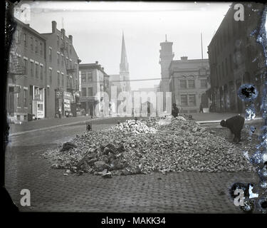 L'horizontale, 1970 Photographie noir et blanc montrant un grand tas de pierres cassées sur une rue pavée. Il y a un homme au travail sur le côté droit de la photo, et un autre homme assis et regardant le spectateur de l'arrière de la pile de gravats. Dans la distance il y a des piétons et du trafic de wagons et une église avec un clocher visible. Titre : les travailleurs de rue à côté d'un tas de gravats sur une rue pavée. . Vers 1910. Holt, Charles Clement, 1866-1925 Banque D'Images