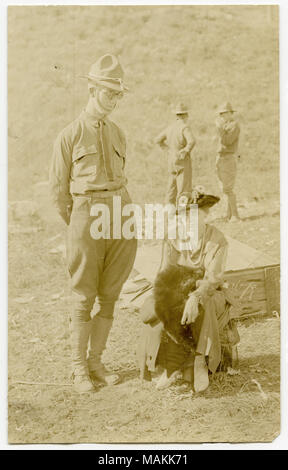 Photographie sépia, verticale montrant un soldat en uniforme debout à côté d'une femme en civil assis sur un objet près du sol dans un champ d'herbe. Deux soldats peuvent être vus en conversation et en face d'une colline en arrière-plan de l'image. Titre : un soldat debout à côté d'une femme assise, tandis que deux soldats converser dans l'arrière-plan. . Entre vers 1914 et vers 1918. Michel, Carl Banque D'Images