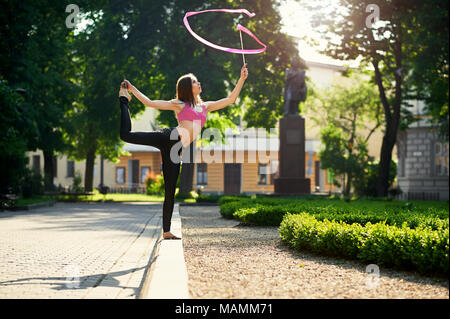 Ivano-Frankivsk, Ukraine - 1 juin 2015 : Jeune fille avec une belle figure est danse avec ruban rouge dans le parc de la ville en matinée. Femme debout sur lit de jardin près de green des semis. Banque D'Images