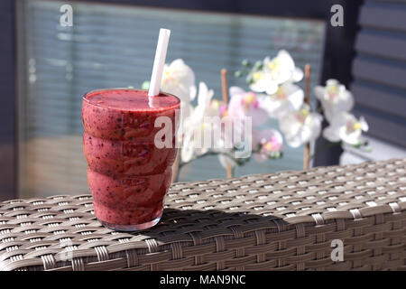 Un verre de smoothie aux fruits rouges fait maison photographiée sur mon balcon pendant une journée d'été. Jolies fleurs blanches et roses phalaenopsis​ sur l'arrière-plan. Banque D'Images