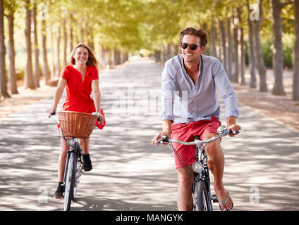 Les jeunes adultes blanc couple riding bikes sur une route bordée d'arbres Banque D'Images