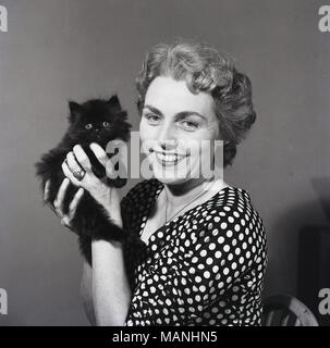 Années 1950, historiques, une dame souriante portant une robe dot poka, contient jusqu'à ses mains son animal de compagnie aimé, un petit mignon chaton poilu, England, UK. Banque D'Images