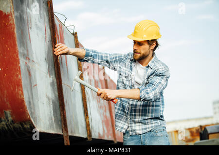 Homme fort avec barbe en chemise à carreaux, très concentré, martelage ou essayer de réparer certains objets rouillés qui exigent de l'attention, à l'extérieur sur un site Banque D'Images