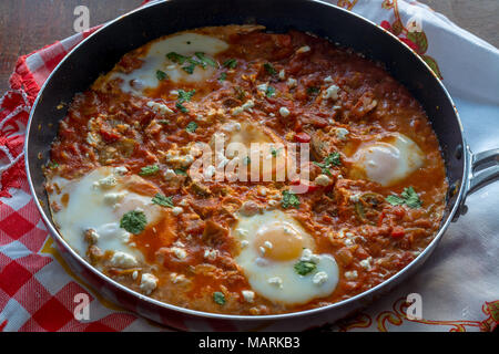 Savoureux et sain Shakshuka, oeufs pochés dans une sauce tomate épicée , poivre dans une poêle, sur table rouge et blanc tissu sur table en bois Banque D'Images