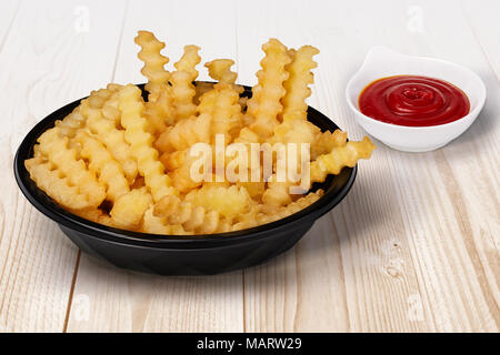 Gros plan de frites Coupe ondulée dans le bol et élimination noir Ketchup Sauce tomate sur le côté sur la table en bois de couleur claire R Fast Food Banque D'Images