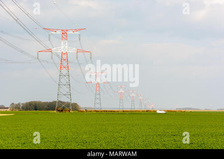 Une ligne électrique aérienne de tension soutenue par les tours de transmission rouge et blanc passant sur un champ cultivé et une route de campagne avec une voiture. Banque D'Images