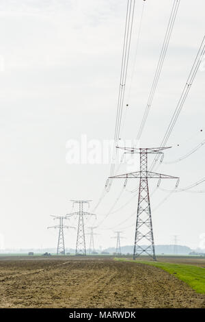 Une rangée de pylônes à haute tension à l'appui d'une ligne électrique aérienne à travers la campagne française, passant sur terre labourée en vertu d'une pâle lumière. Banque D'Images