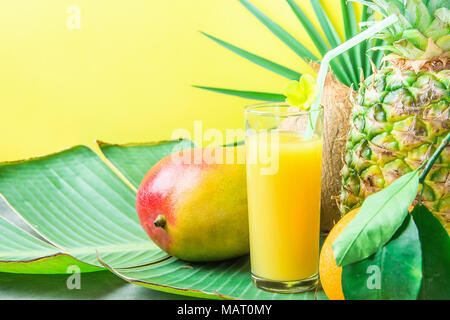 Composition Grand Verre à jus de fruits tropicaux fraîchement pressé avec de la paille de coco ananas Mangue Orange sur vert grande feuille de palmier. La lumière du soleil. L'été Banque D'Images