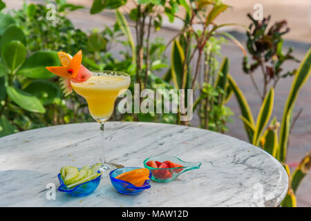 Verre à cocktail jaune en faveur avec morceau de pastèque sur le dessus et entouré de bols de tranches de fraise, citron, orange sur table en marbre ronde Banque D'Images