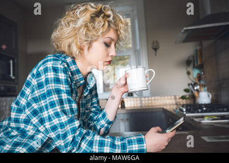 Woman having coffee lors de l'utilisation de téléphone mobile dans la cuisine Banque D'Images