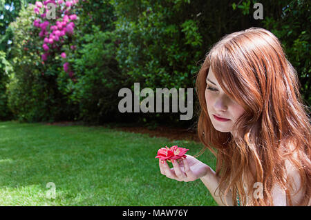 Une femme aux cheveux rouges assis dans un jardin tenant une fleur. Banque D'Images