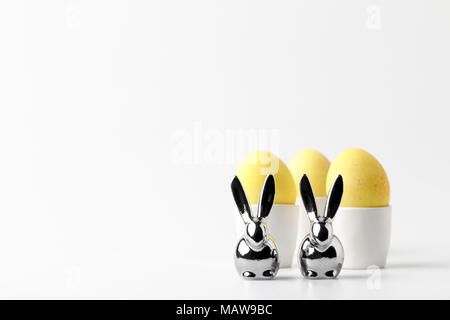 Les oeufs de Pâques peints en jaune dans l'oeuf se tient et des statuettes de lapins on white Banque D'Images