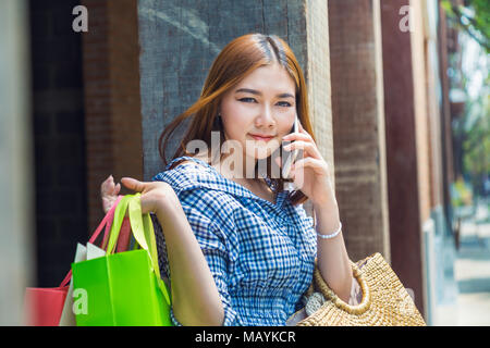 Young Asian woman carrying shopping bags son par dessus son épaule et en utilisant son téléphone à l'extérieur du shopping plaza, bon pour le shopping ou yo Banque D'Images