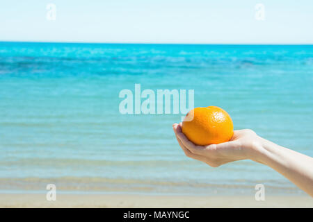 Young Caucasian Female Hand Holding ensemble orange mûre sur Ciel Bleu mer Turquoise Background. Vagues de sable blanc. Les vacances d'été Wanderlust voyageant Con Banque D'Images