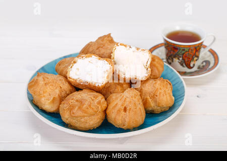 Frais Profiteroles profiteroles gâteaux remplis de crème pâtissière dans une assiette blanche et d'une tasse de thé avec paisley design sur fond de bois blanc de la population. Banque D'Images
