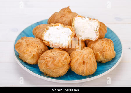Frais Profiteroles profiteroles gâteaux remplis de crème pâtissière et avec l'un coupé en deux dans une plaque bleu turquoise sur fond blanc. populaires Persi Banque D'Images