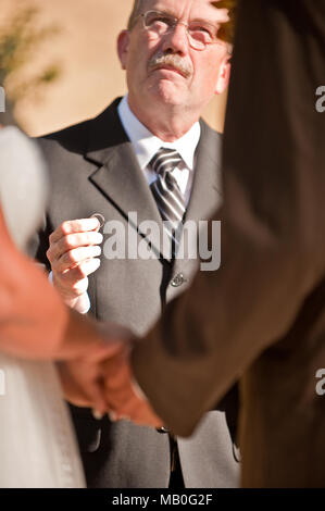 Un prêtre officie un mariage avec une bague de mariage dans la main en face de jeunes mariés qui tiennent par la main lors d'une cérémonie de mariage en plein air Banque D'Images