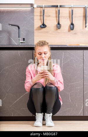 Mode jeune femme sérieuse en utilisant son téléphone portable dans la cuisine comme elle s'accroupit vers le bas appuyé contre un cabinet Banque D'Images