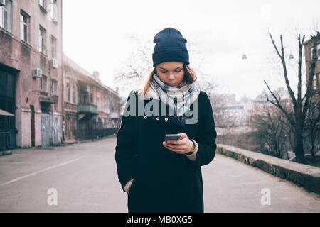 Vue avant du jeune femme au centre de la rue la marche et regarde dans téléphone intelligent. Habillé à la mode fille dans un manteau noir, écharpe bleue et hat w Banque D'Images