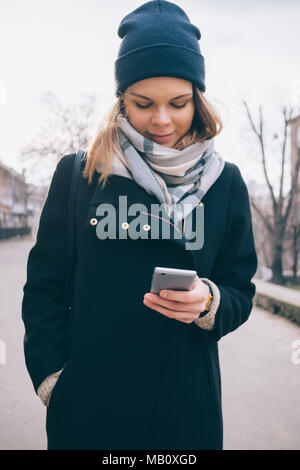 Vue de face souriante jeune femme dans la rue et permanent à la recherche de téléphone intelligent. Girl wearing élégant manteau noir, écharpe et chapeau Bonnet bleu dans Win Banque D'Images