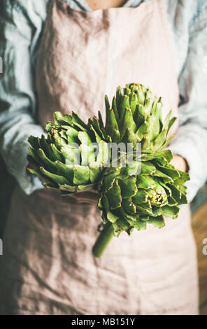 La productrice de porter un tablier de lin pastel et shirt holding artichauts frais dans ses mains, selective focus, composition verticale. Les produits biologiques ou lo Banque D'Images