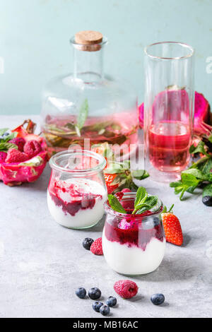 Pots de yaourt naturel blanc avec sauce aux fruits, salade de fruits avec des fruits du dragon rose, baies et menthe, servi avec une bouteille de limonade sur la table gris. Hea Banque D'Images