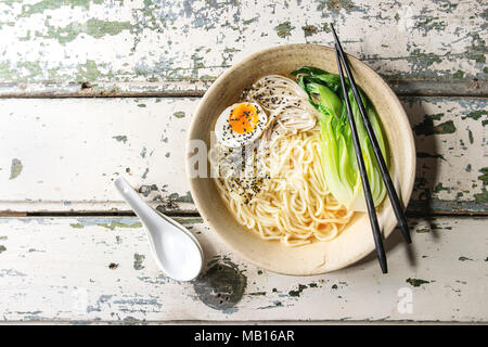 Plat asiatique nouilles Udon avec oeuf mollet, sésame, champignons, boc choy servi dans un bol en céramique avec cuillère et baguettes sur fond de bois blanc Banque D'Images