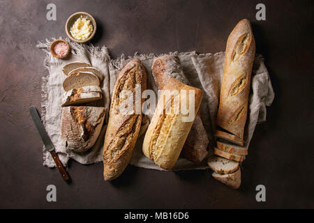 Variété de pâtisserie frais loafs seigle artisan, blanc et le pain de grains entiers sur tissu en lin avec du beurre, sel et rose vintage couteau sur la texture marron foncé Banque D'Images