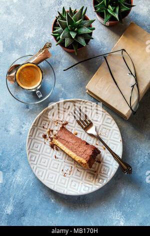 Une tranche de gâteau au fromage au chocolat sur une assiette, tasse de café, vieux livre, verres et plantes succulentes sur arrière-plan Banque D'Images