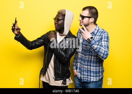 Deux meilleurs amis multiraciale en tenant l'appareil photo de téléphone mobile avec selfies isolé sur fond jaune. Banque D'Images