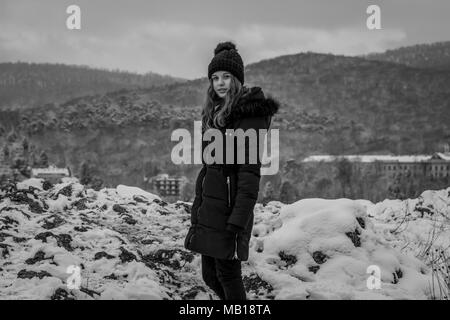 Jolie teen girl in snow, face caméra, noir et blanc. Belle Caucasian teenage girl in manteau noir, les montagnes enneigées en arrière-plan. Banque D'Images