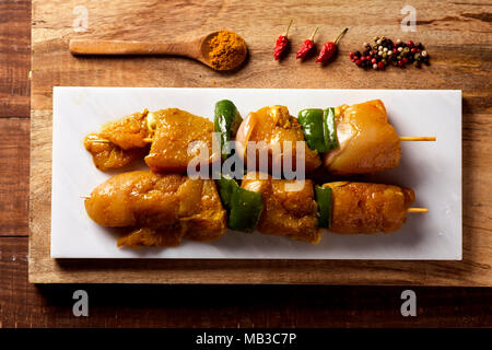Portrait de quelques brochettes de poulet cru épicé sur une plaque en céramique blanc placé sur une table en bois, à côté de quelques épices, comme le curry, Banque D'Images