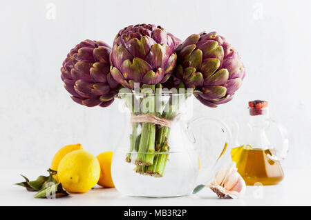 Des tas d'artichauts violets dans un décanteur avec de l'eau sur fond blanc avec du citron, l'huile d'olive , ail . printemps alimentation saine Banque D'Images