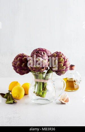 Des tas d'artichauts violets dans un décanteur avec de l'eau sur fond blanc avec du citron, l'huile d'olive , ail . printemps alimentation saine Banque D'Images