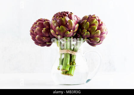 Des tas d'artichauts violets dans un décanteur avec de l'eau sur fond blanc . printemps alimentation saine with copy space Banque D'Images