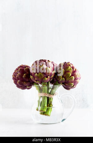 Des tas d'artichauts violets dans un décanteur avec de l'eau sur fond blanc . printemps alimentation saine with copy space Banque D'Images